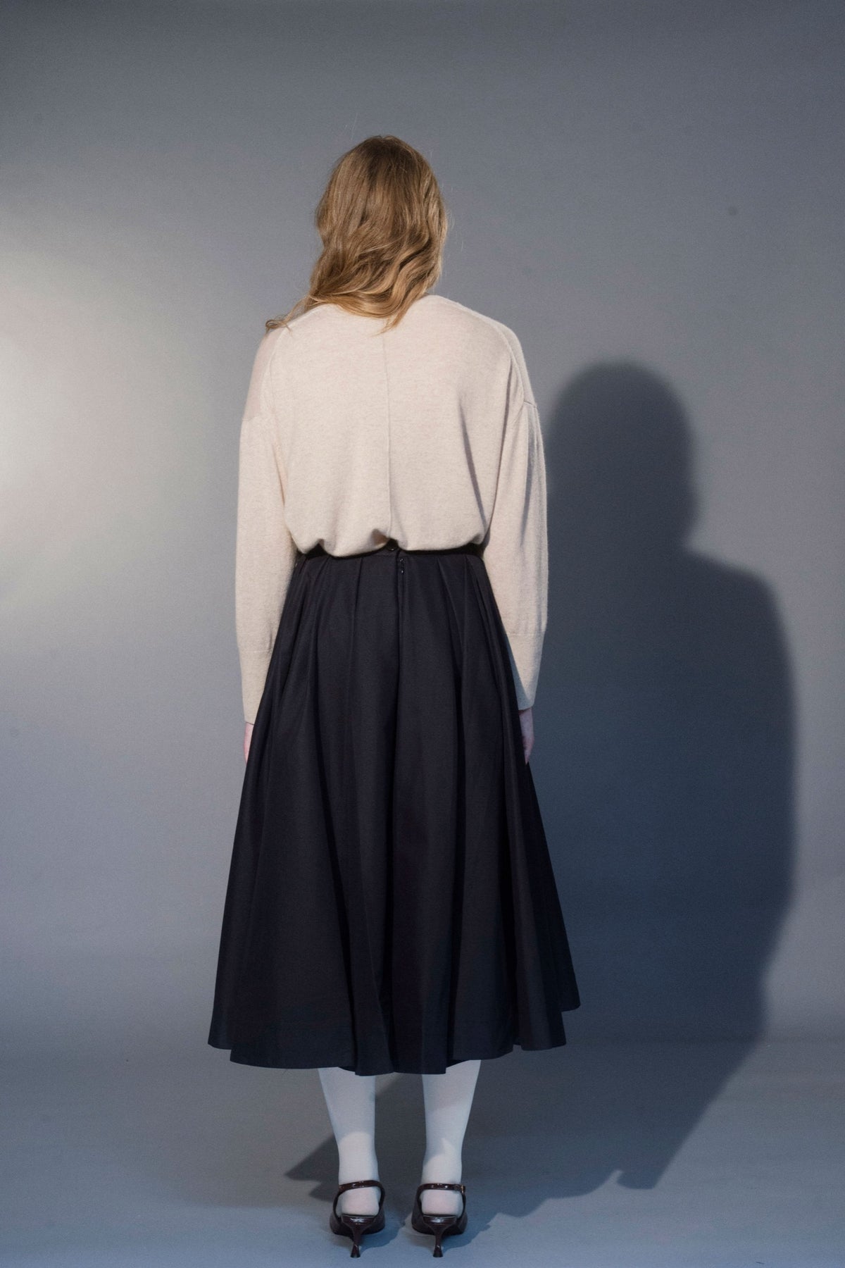 Woman wearing the Stathe Vinorama Skirt in Black with the Salvia V-neck Sweater — editorial, back view